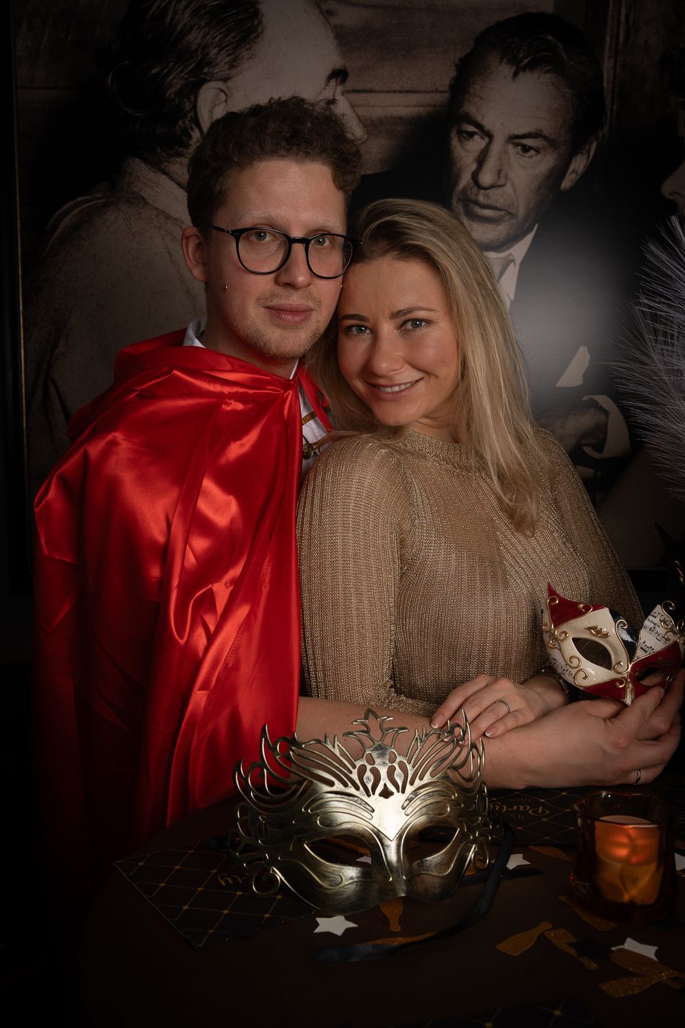 A couple poses for a romantic masquerade-themed photo with decorative masks and a red cape in dimly lit setting.
