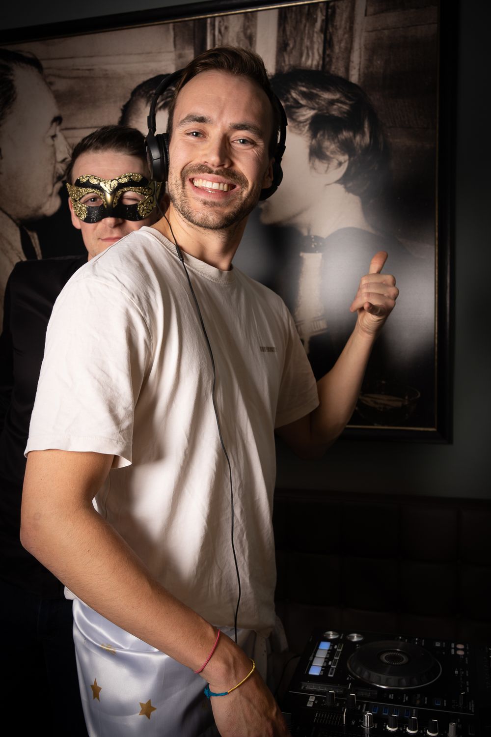 A DJ in a white t-shirt smiles at a party with DJ equipment visible and someone wearing a mask in the background.
