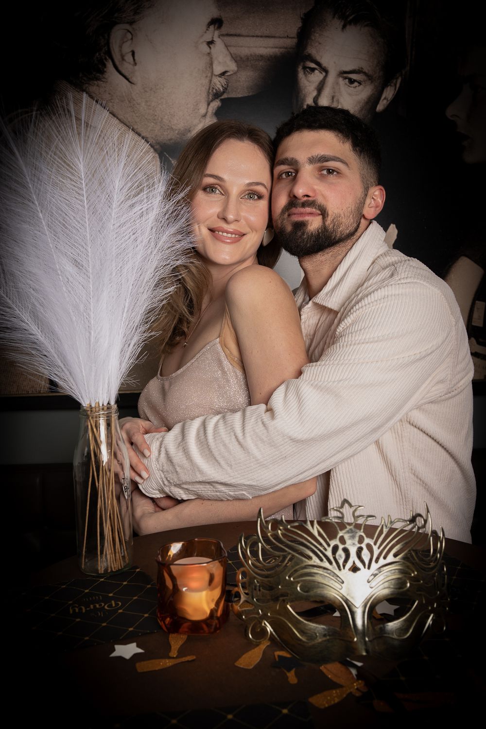 A couple in light colored clothes poses by a table with masquerade masks and feathers in romantic lighting.