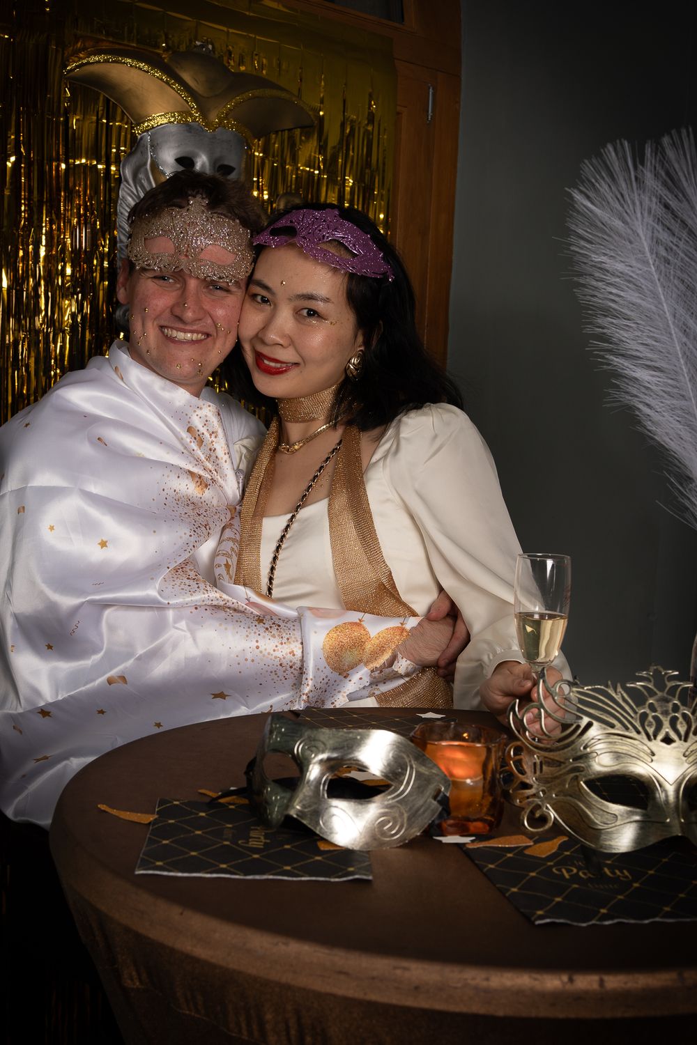 A couple in white attire enjoys champagne at a masquerade party with gold decorations and masks on the table.