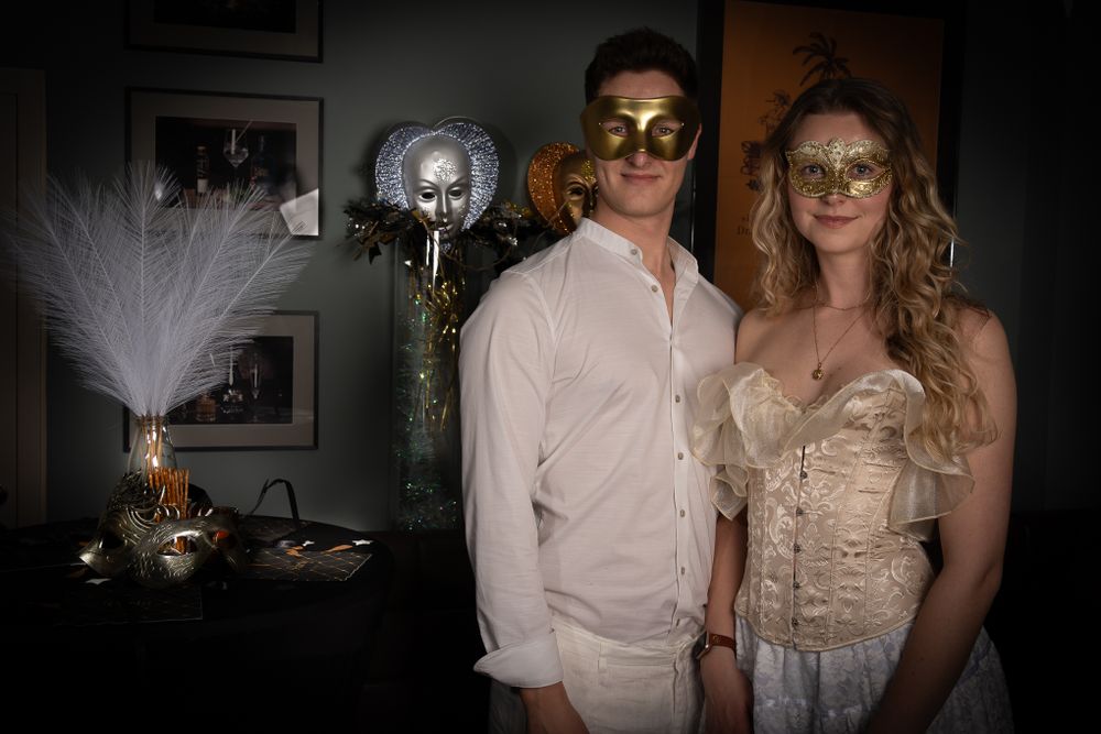 A couple poses in white formal attire and ornate gold masks at a masquerade party with decorative feathers in background.