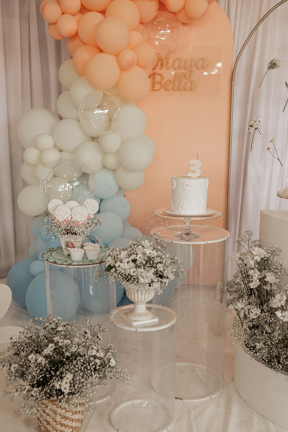 Elegant party setup with peach and blue balloon arch, tiered cake display stands, and baby's breath floral arrangements.