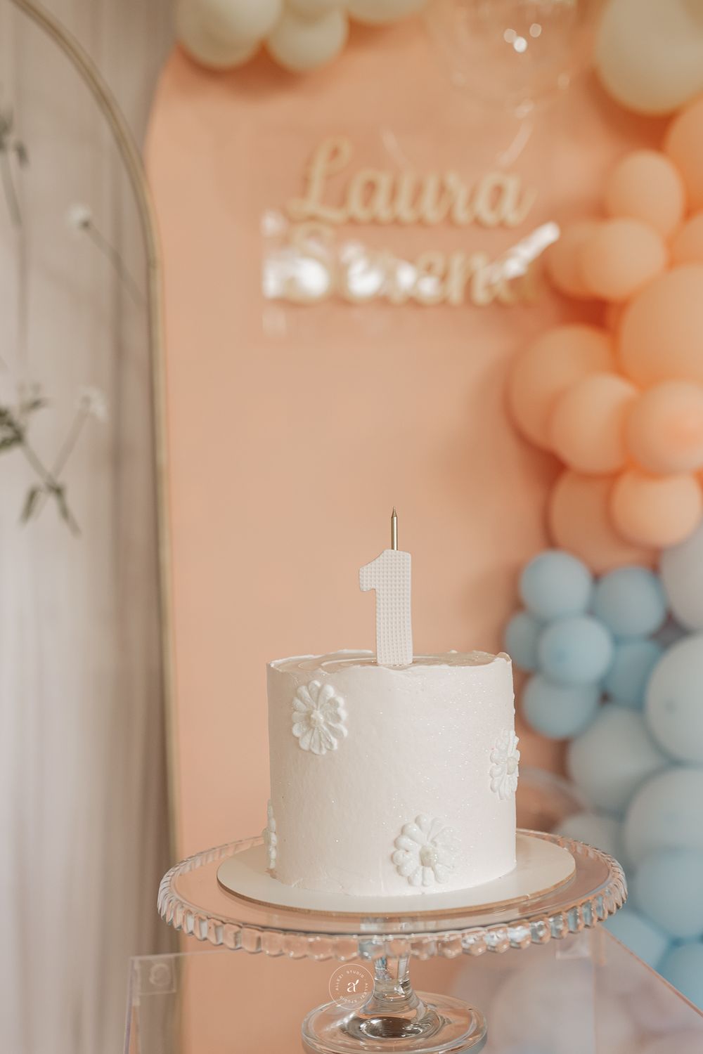 A simple white first birthday cake with floral details sits on a crystal stand against peach and blue balloon decorations.
