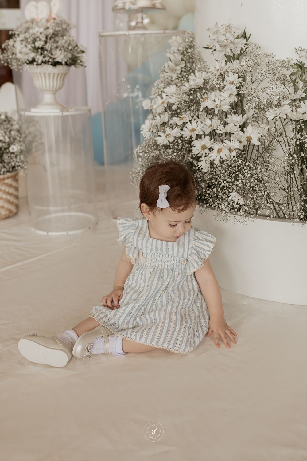 A small child wearing a gray striped dress with ruffled sleeves sits on a light floor near white floral decorations.