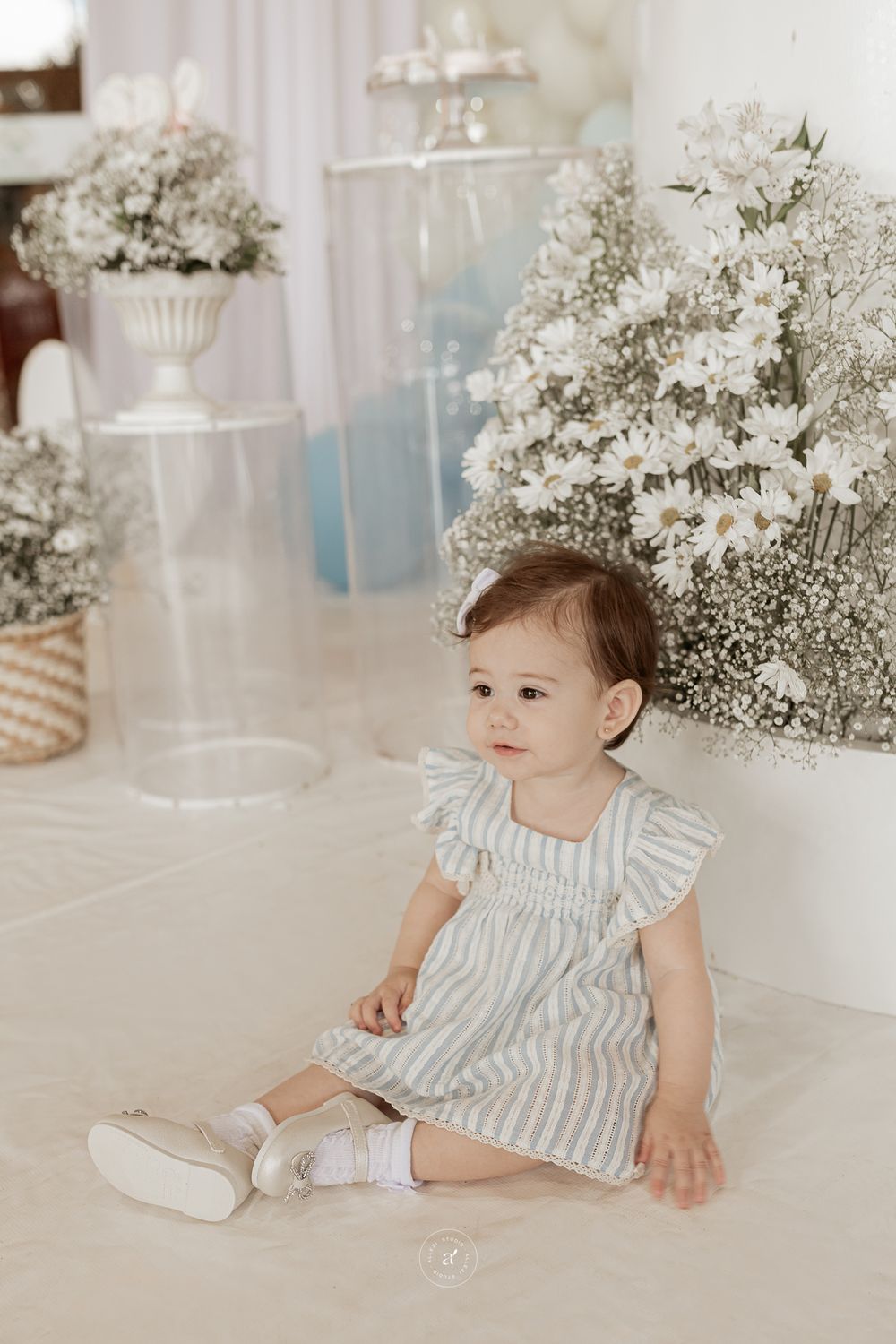 A small child in a striped dress sits on a white set with baby's breath floral arrangements in the background.