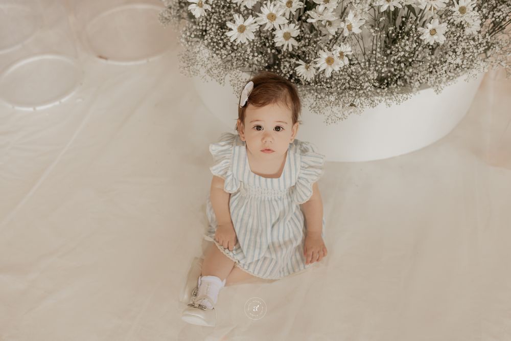 Baby wearing white ruffled dress sits on beige backdrop with dreamy floral decor overhead.