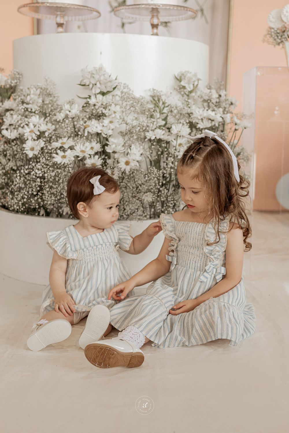 Two children in matching white ruffled dresses sit together against a backdrop of baby's breath flowers.