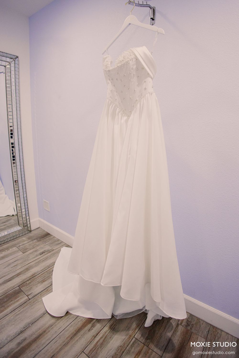 Full-length white wedding gown displayed on a hanger against a pale blue wall with hardwood flooring.