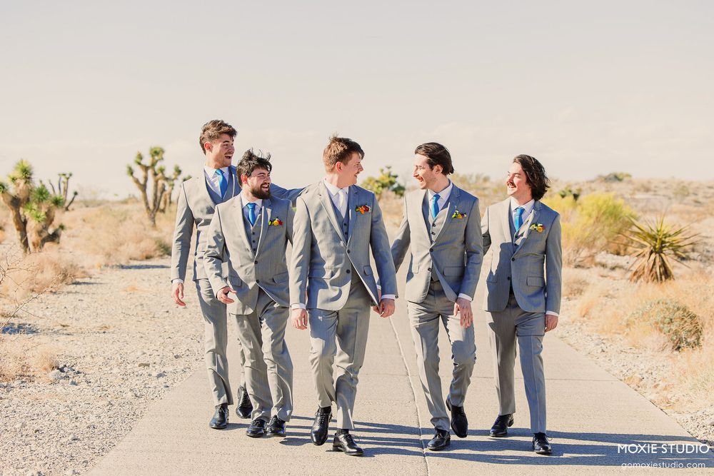 Groomsmen in gray suits walk together on a desert road with Joshua trees in the background during sunset.