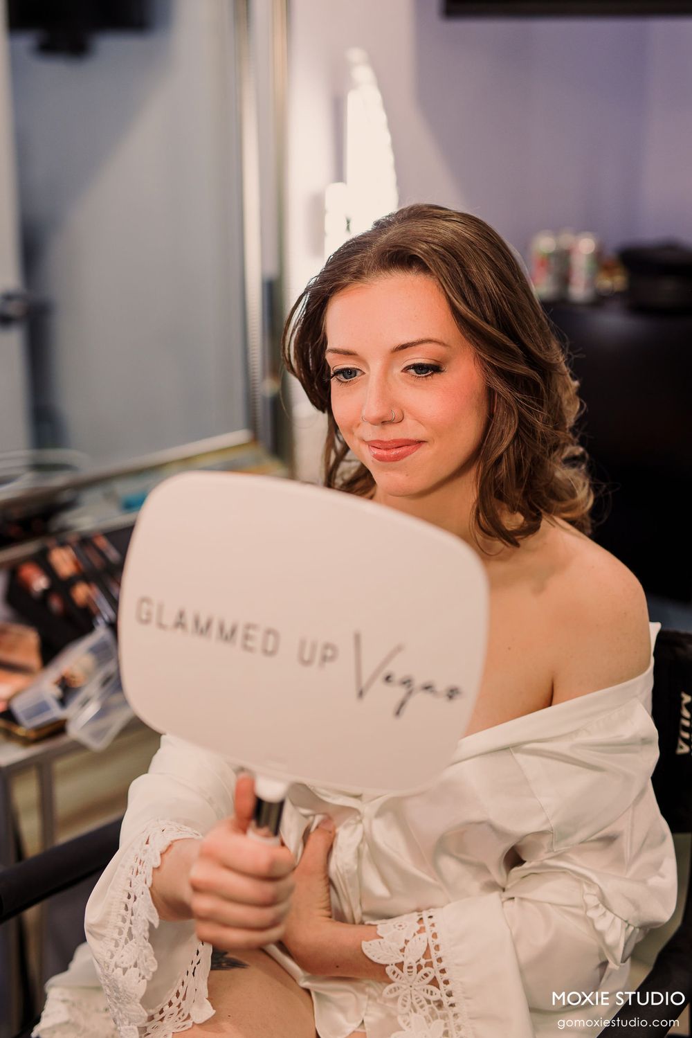 A woman in a white lace robe holds a makeup mirror during wedding preparations.