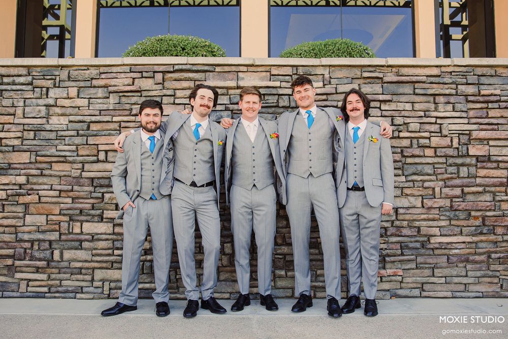 Groomsmen in matching gray suits pose together against a stone wall exterior.