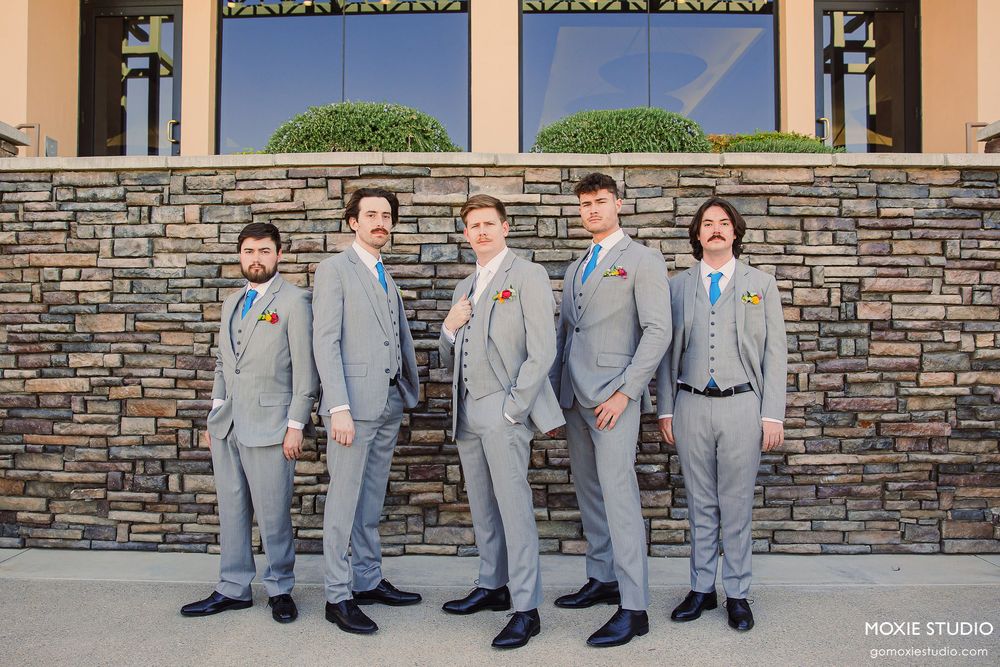 A group of groomsmen in matching light gray suits pose together in front of a stone wall on a sunny day.