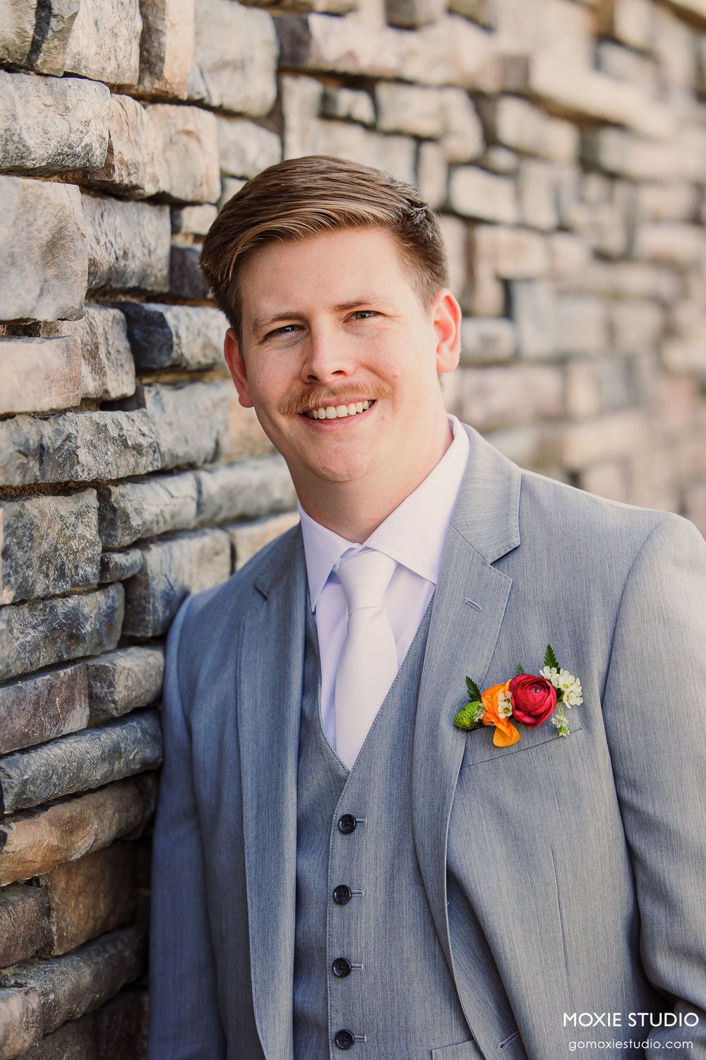 Sequence of portrait shots showing person in light gray suit with colorful boutonniere against stone wall.