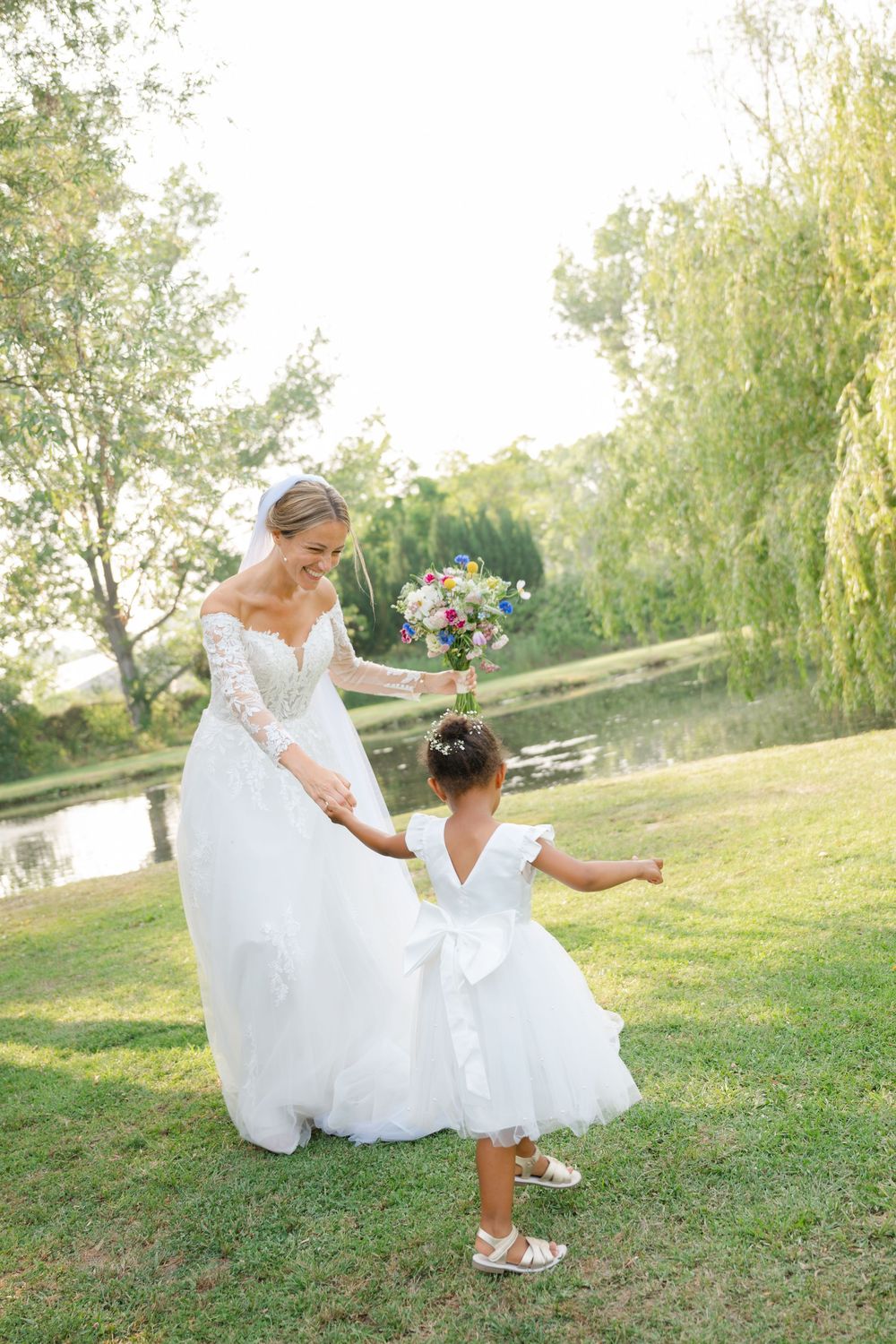 Bride in flowing white dress sits playfully on grassy hillside on sunny day at outdoor venue.