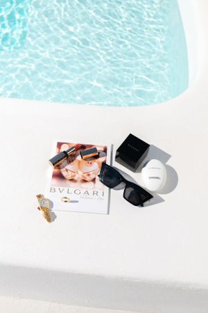 Luxury accessories including sunglasses and jewelry arranged by poolside with fashion magazine.