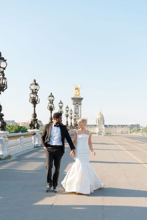 A couple in wedding attire walks across the historic Pont Alexandre III bridge in Paris during sunset.
