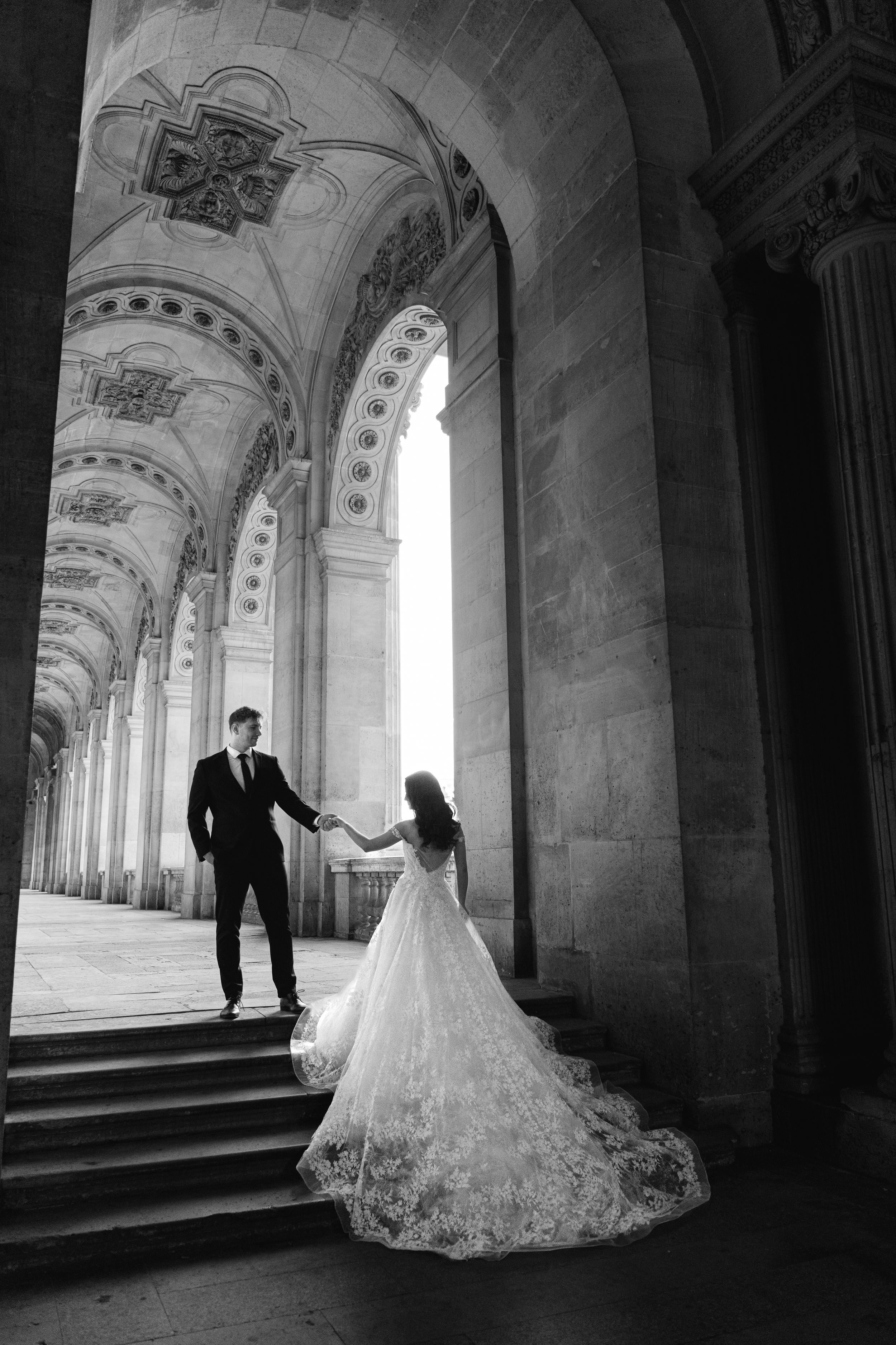 A romantic wedding photoshoot in a grand stone archway corridor with dramatic lighting and shadows.