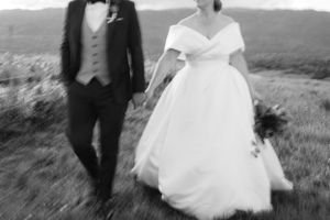 Artistic black and white photo of couple walking through mountain field in wedding attire.