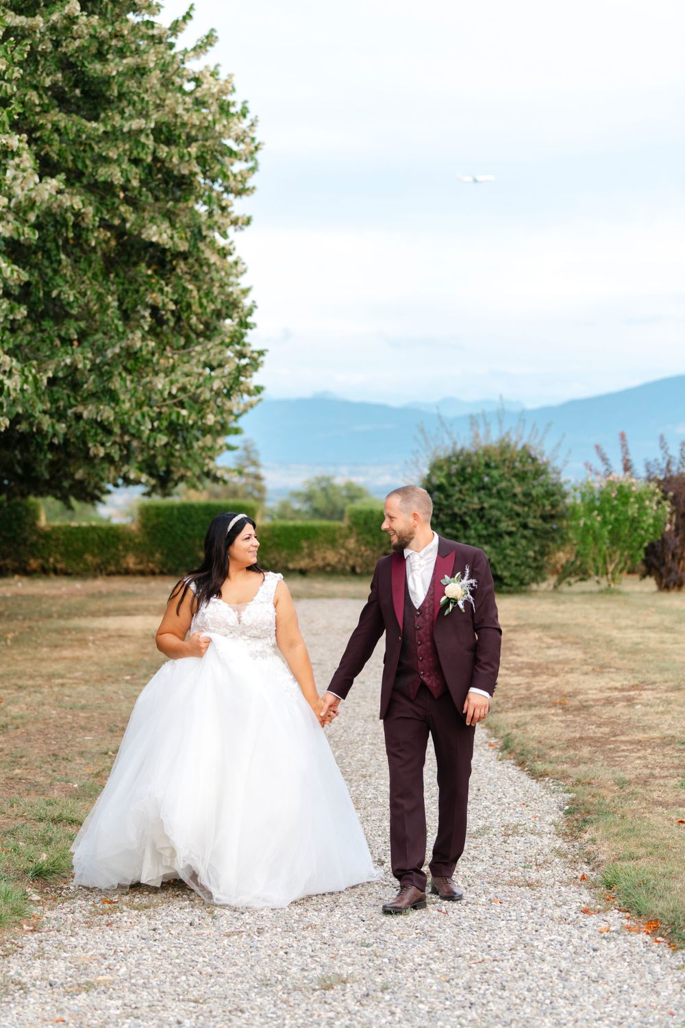 Wedding couple walking hand in hand on gravel path with mountains and blooming trees in background.
