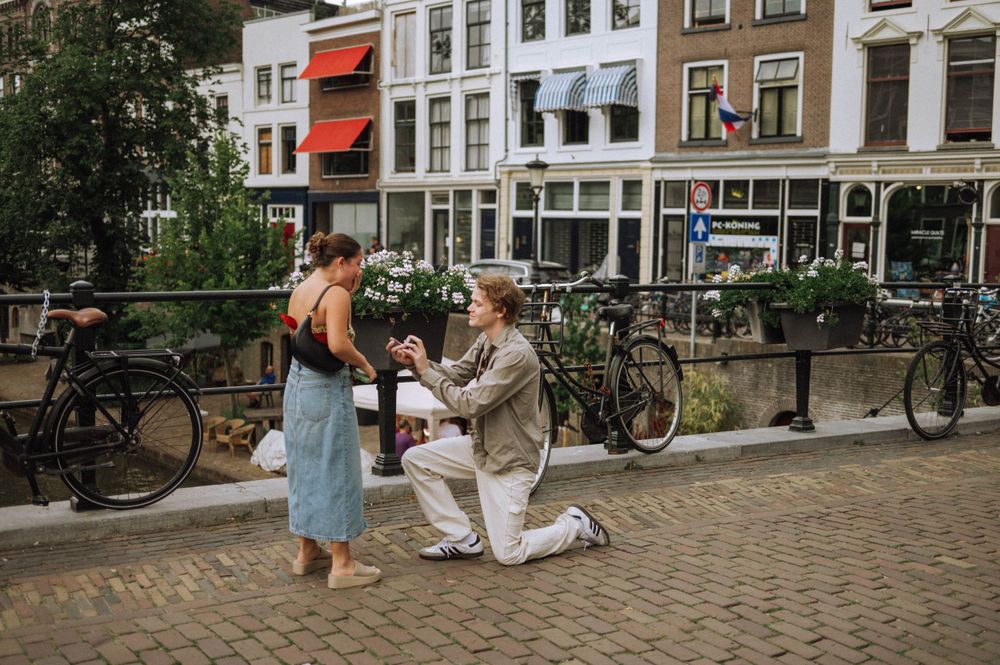A romantic couple shares an intimate moment along a canal in a European city with historic buildings in the background.