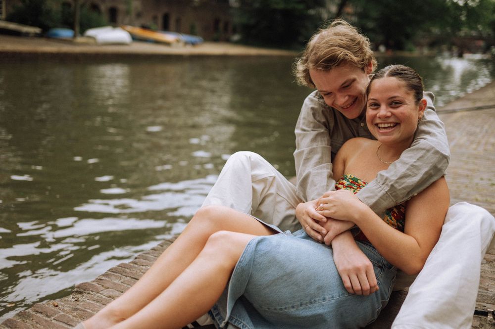 A couple shares a relaxed moment by the water's edge in a European city setting.