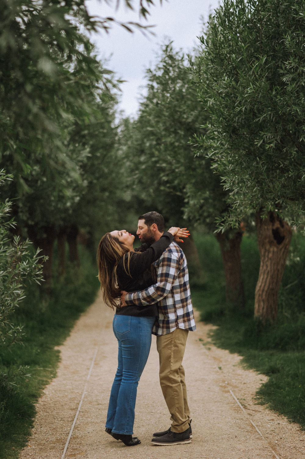 A couple embraces romantically on a tree-lined path in a garden setting.