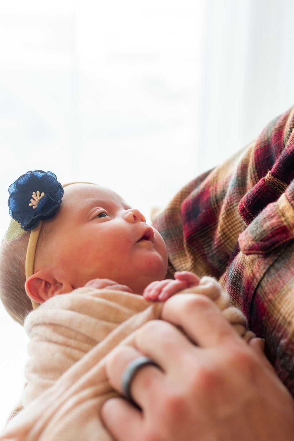Baby wearing a blue flower headband looks up while being cradled in someone's arms wearing a plaid shirt.