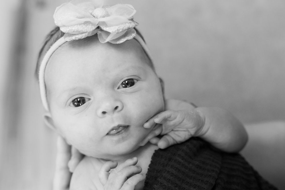 Black and white portrait of a baby wearing a white headband with bow while looking at the camera with curious eyes.