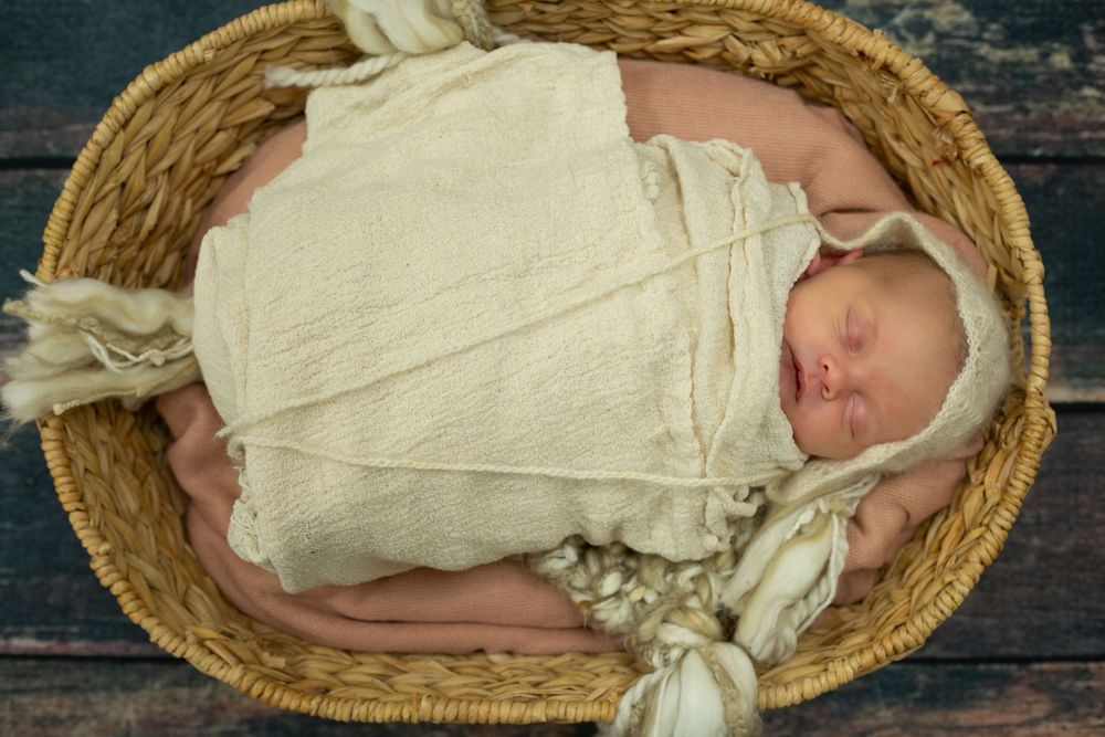 Sleeping newborn wrapped in white swaddle lies peacefully in rustic woven basket on wooden surface.