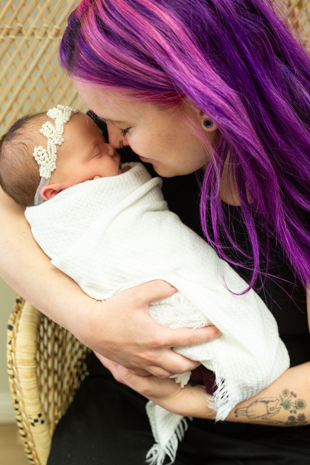 A sleeping baby in a lace headband is held by someone with vibrant purple hair and visible arm tattoos.