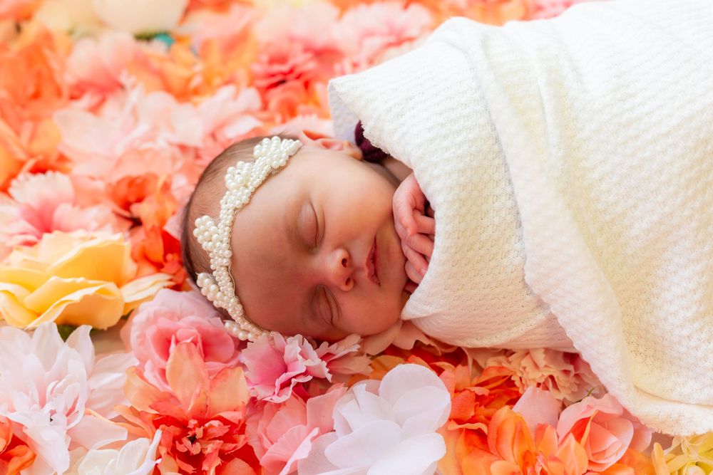 Sweet baby sleeping peacefully on a bed of pink, peach and yellow flowers while wrapped in white blanket.