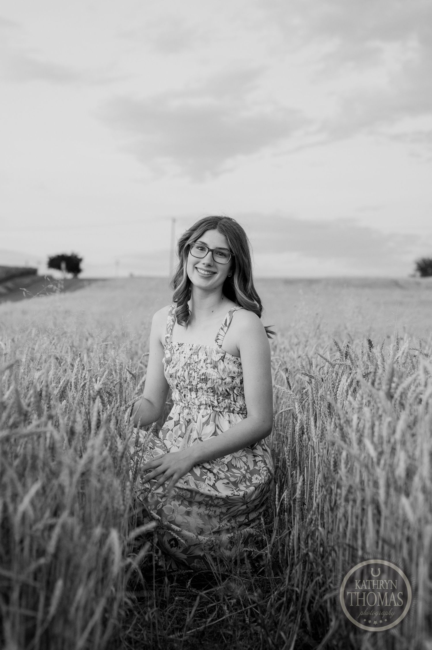 Black and white portrait of someone in floral dress standing among tall wheat stalks.
