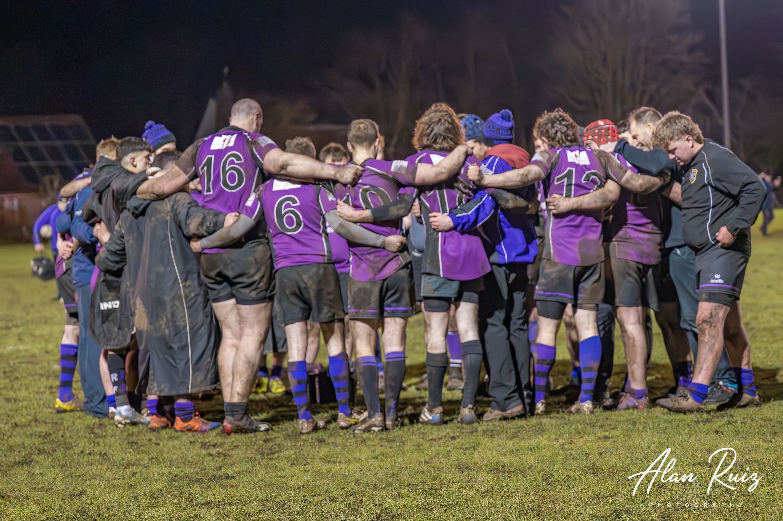 Rugby team members stand together in a huddle formation on a floodlit field at night.