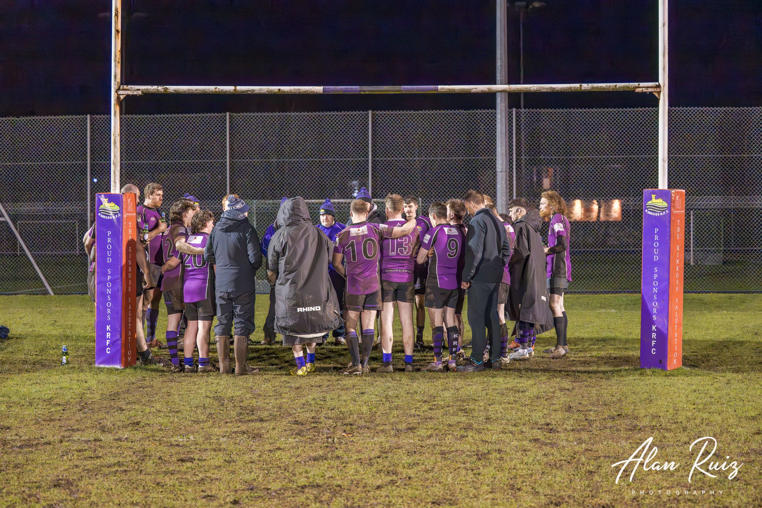 Rugby goal posts stand tall against dark night sky with players gathered in distance.