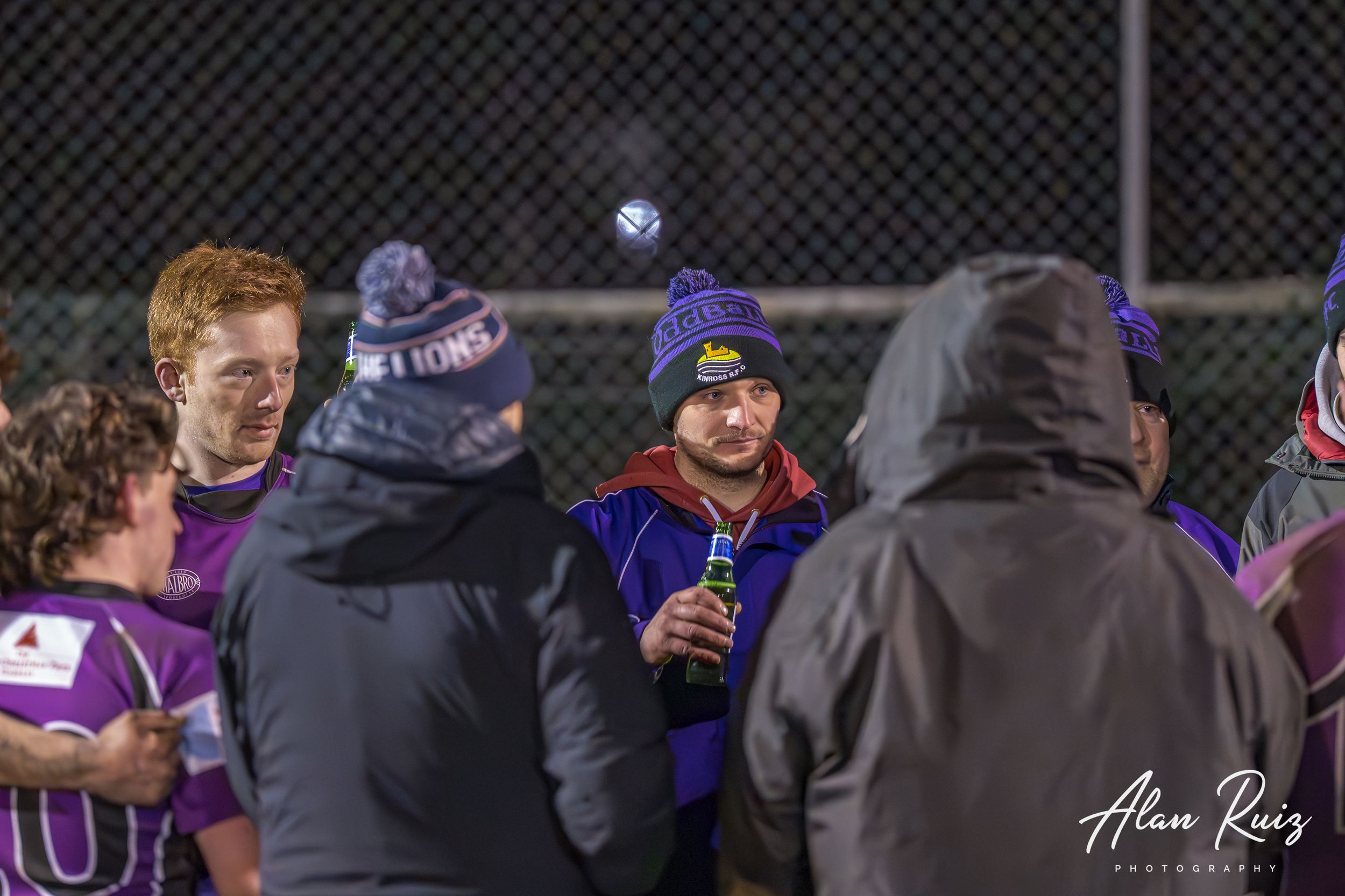 Rugby players gather near chain-link fence under floodlights during evening match.