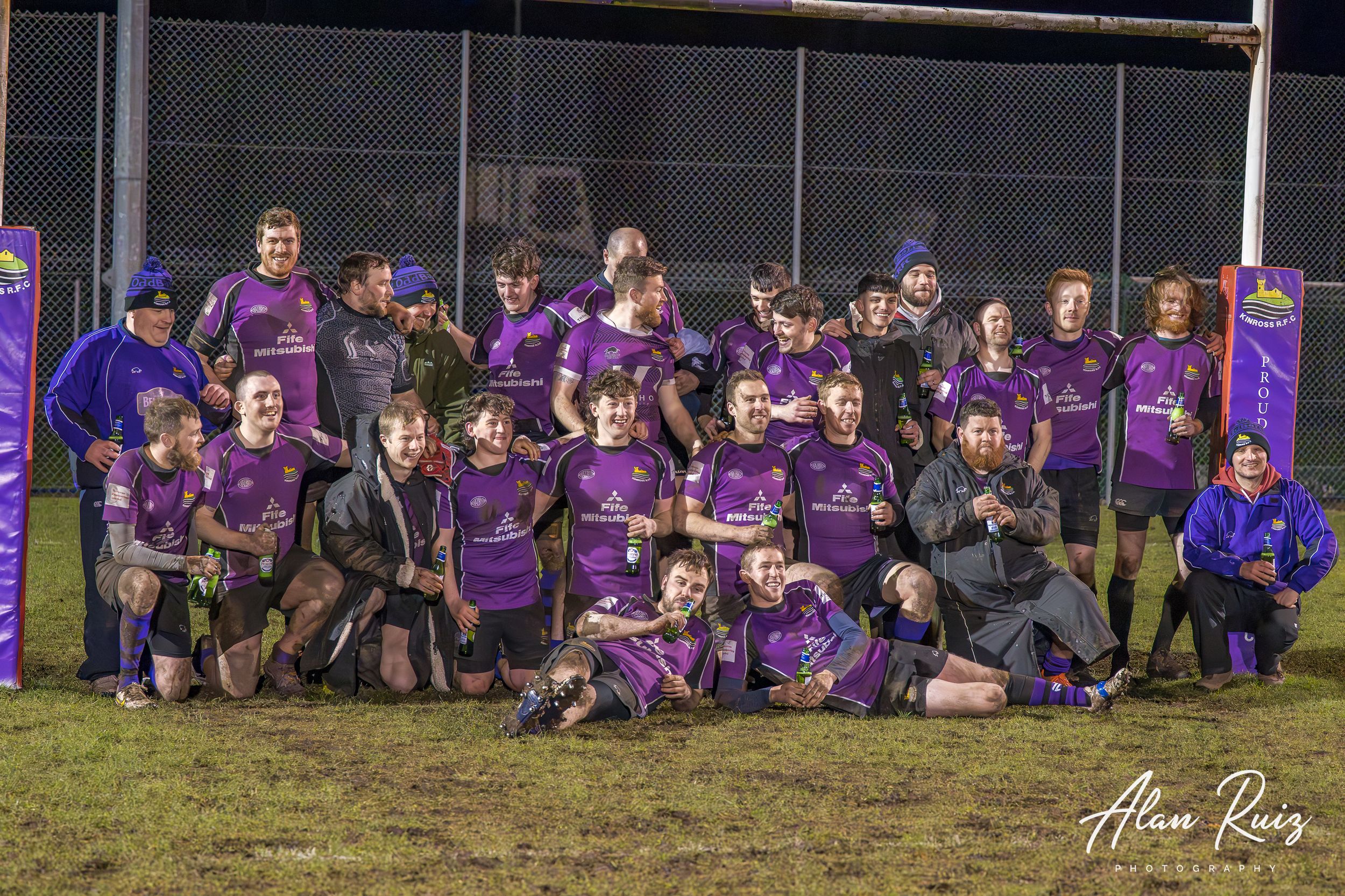 Rugby team in purple uniforms poses for group photo beneath goal posts at night.