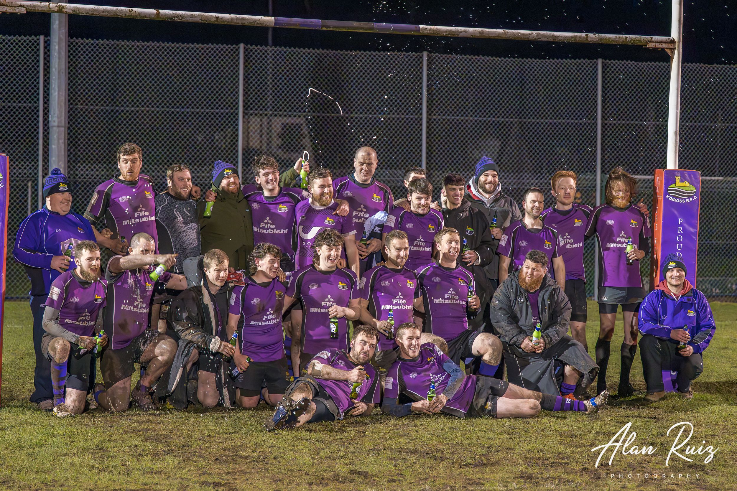 Rugby players in purple jerseys line up for team photo under floodlights on field.