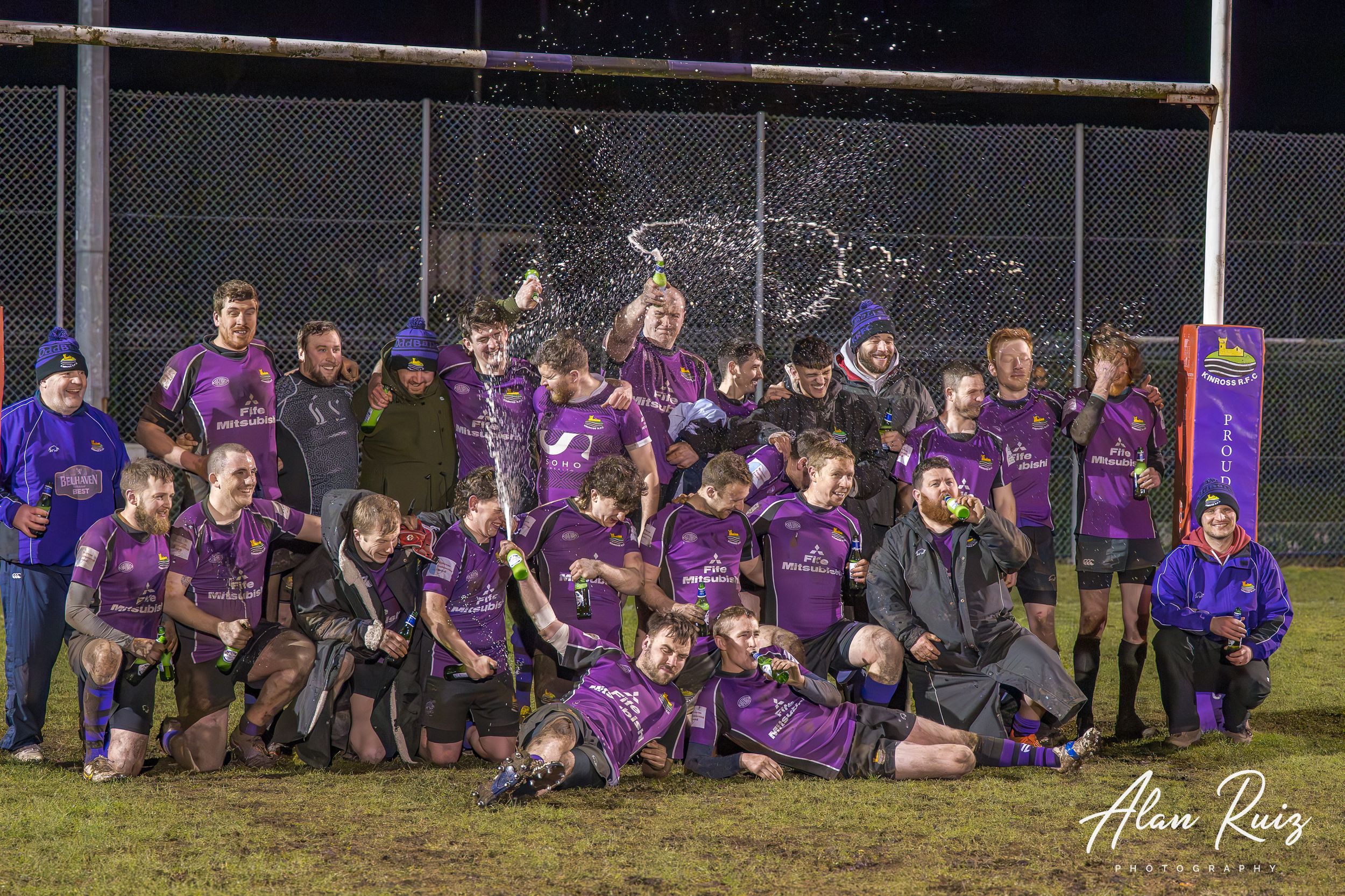 Team photo session of rugby players in purple uniforms on floodlit field at night.