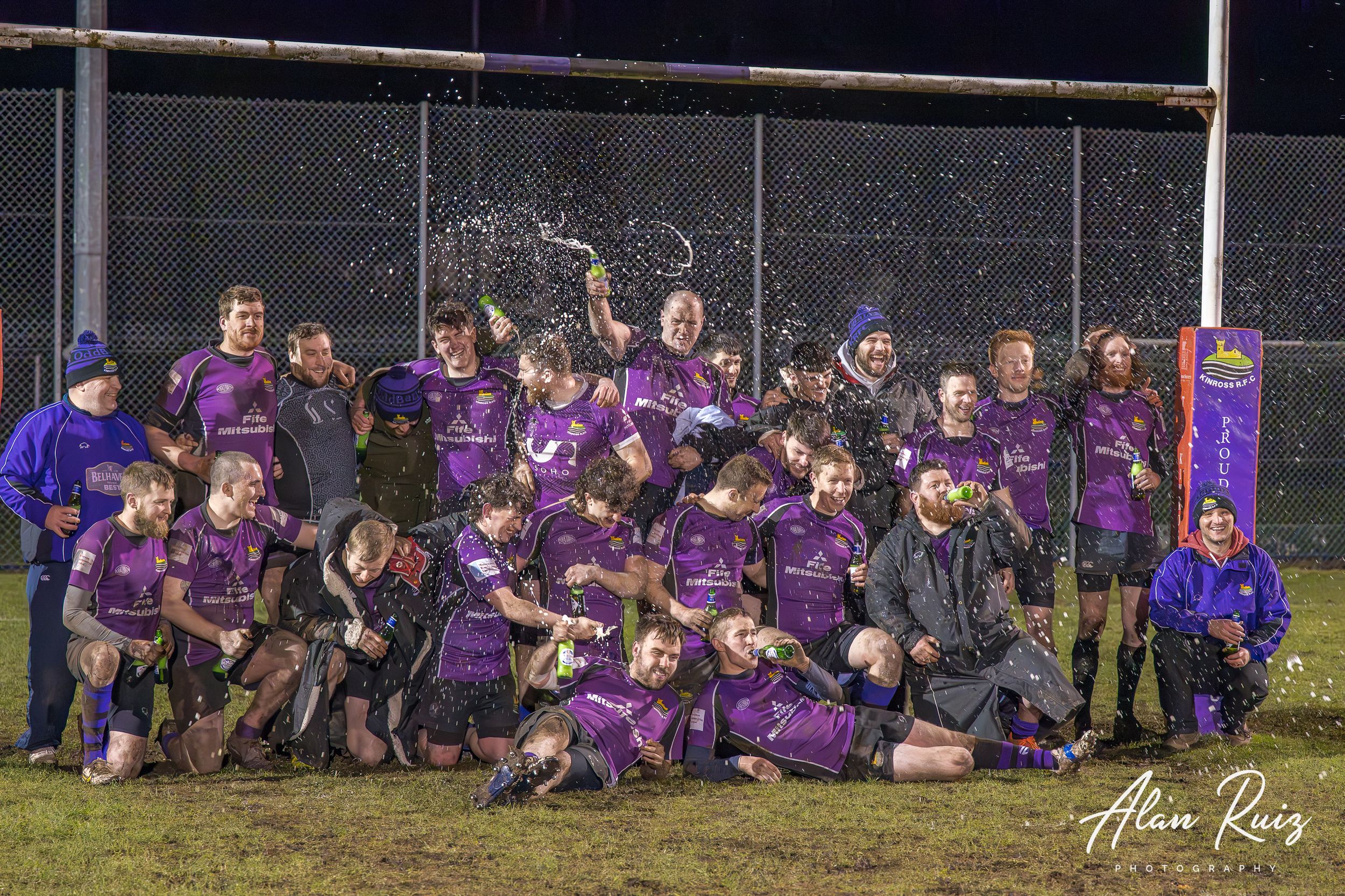 Rugby squad poses together for group photograph on muddy field under night lights.
