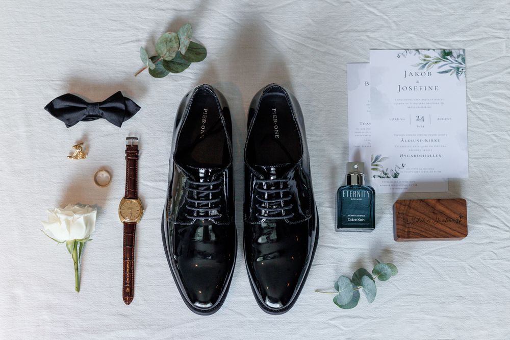 Wedding details flatlay featuring black formal shoes, watch, invitation, white rose, eucalyptus and accessories on white surface.