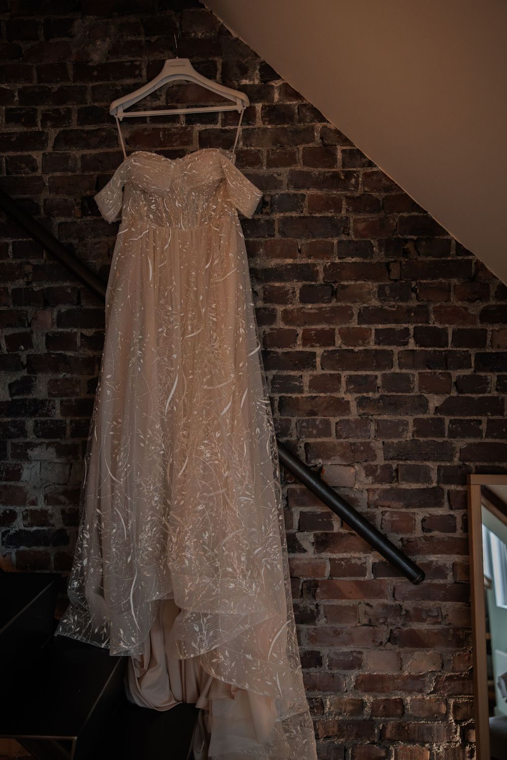 A delicate lace wedding dress with off-shoulder design hangs against an exposed brick wall in dramatic lighting.