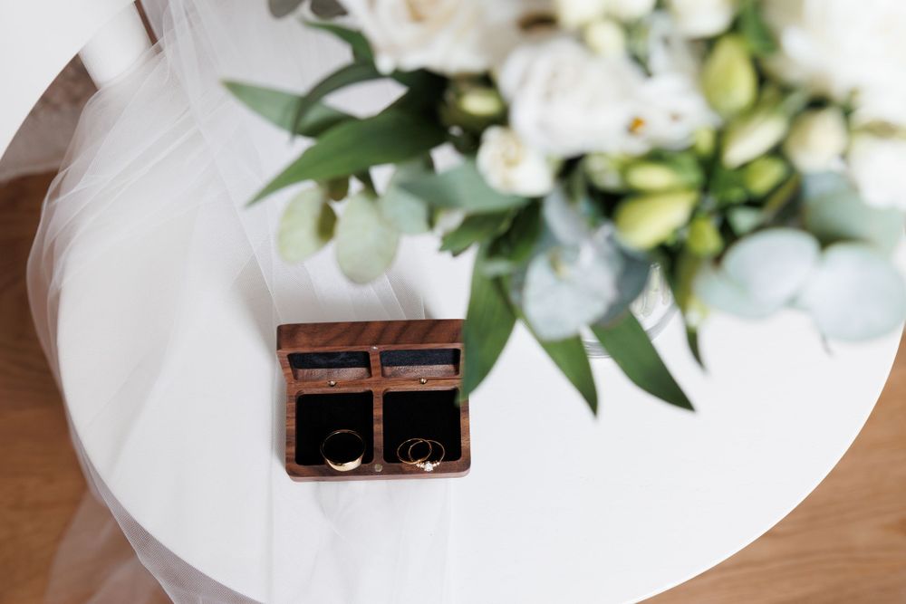 Two wedding rings rest in a small wooden box on a white table with white flowers in soft focus above.