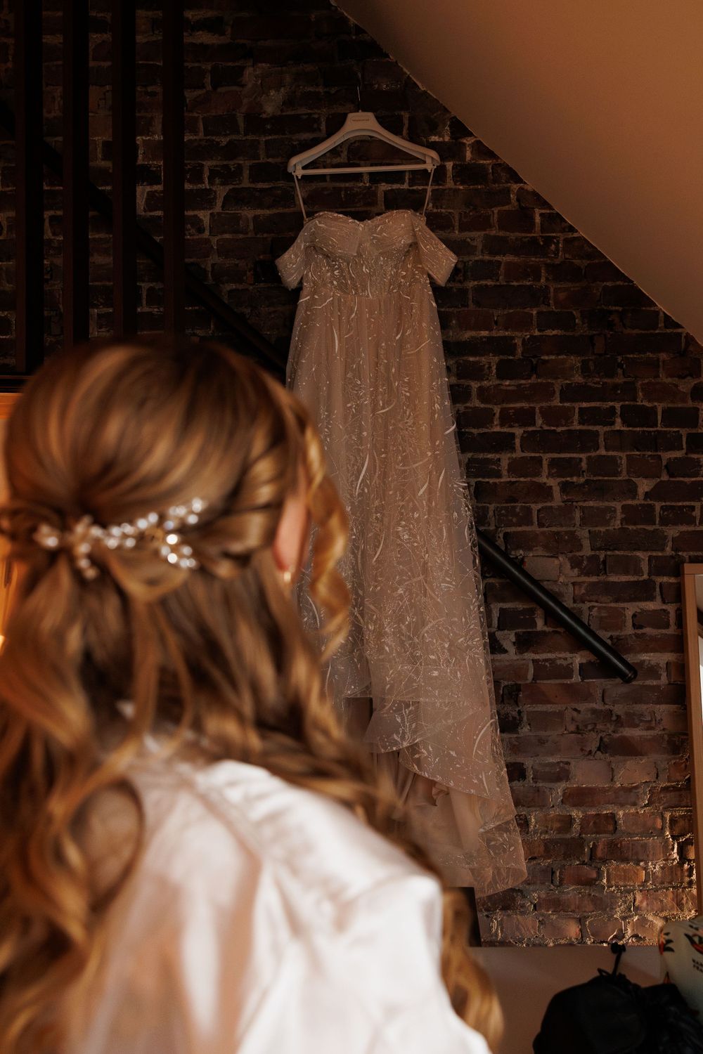 Beautiful beaded wedding gown hangs on display against rustic brick wall while styled hair with floral accessories appears in foreground.