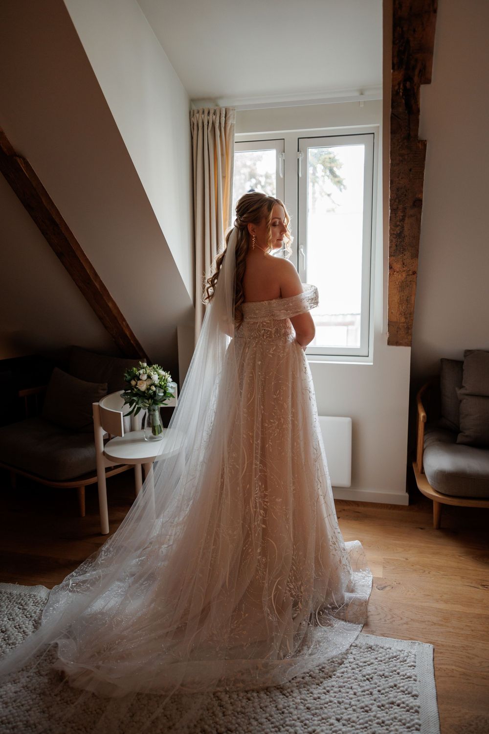A bride in an off-shoulder lace wedding gown with flowing veil stands by a window in a rustic attic room.