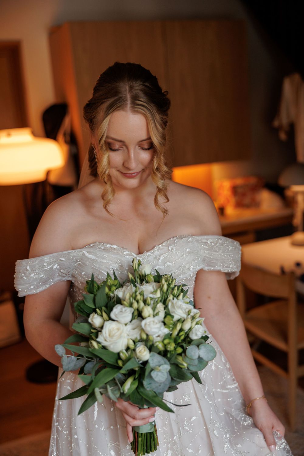 Bride in off-shoulder lace wedding dress holds elegant white and green bouquet in warm-lit interior setting.