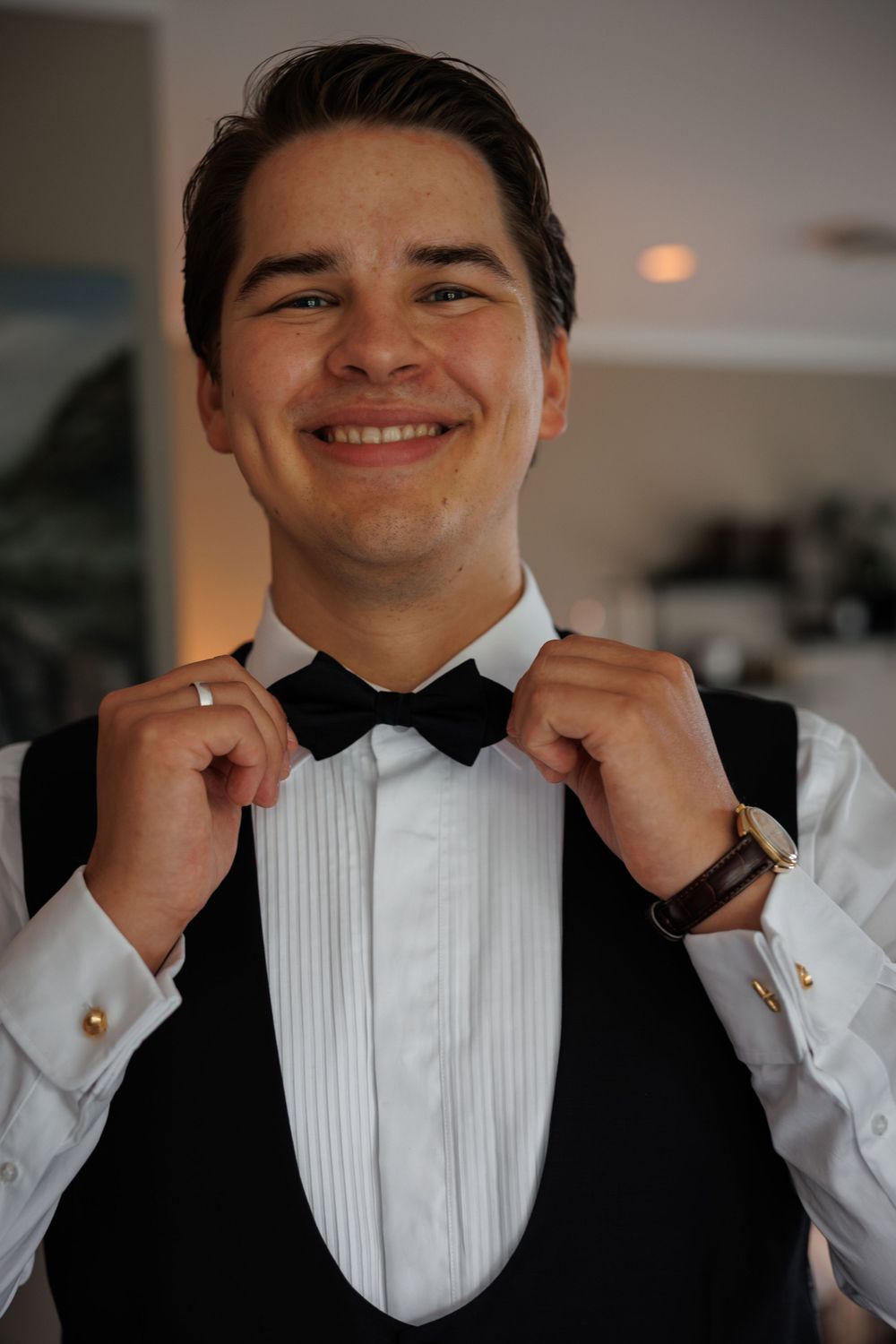 A person in formal attire adjusts their black bow tie while wearing a white dress shirt and vest.