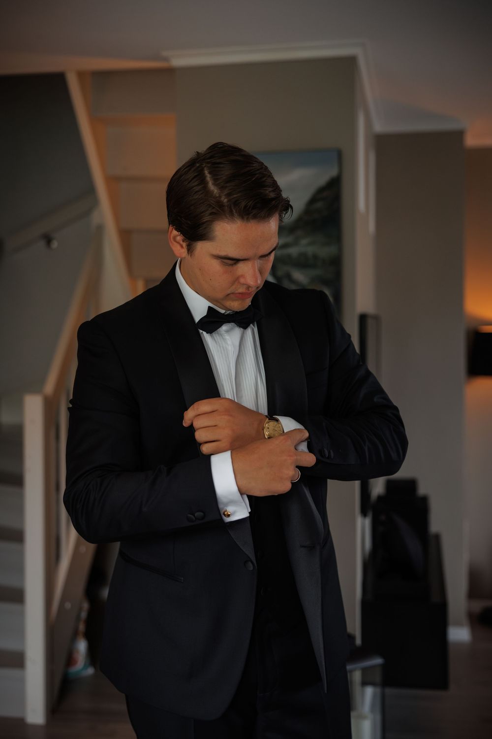 A person in a formal black tuxedo adjusts their sleeve cuff while getting dressed in a dimly lit room.