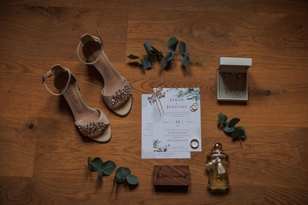 Wedding details flat lay with invitation, beige sandals, eucalyptus leaves and decorative items on wooden surface.