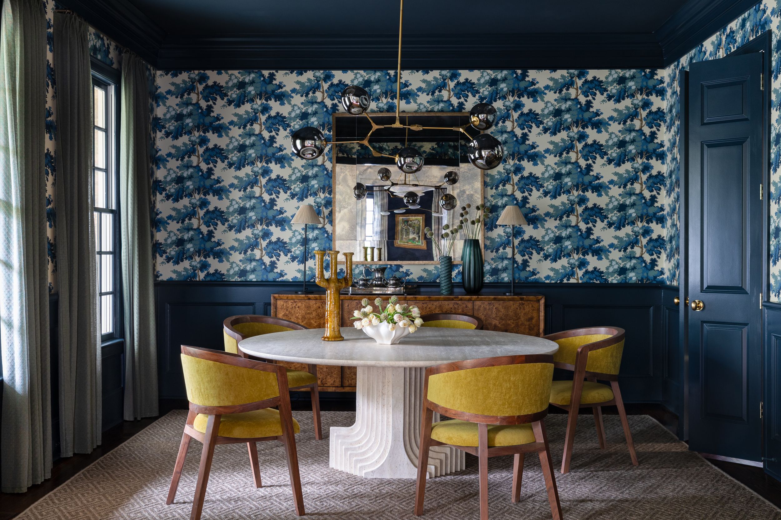 Navy blue floral wallpaper adorns the walls of an eclectic dining room with yellow velvet chairs and modern decor.