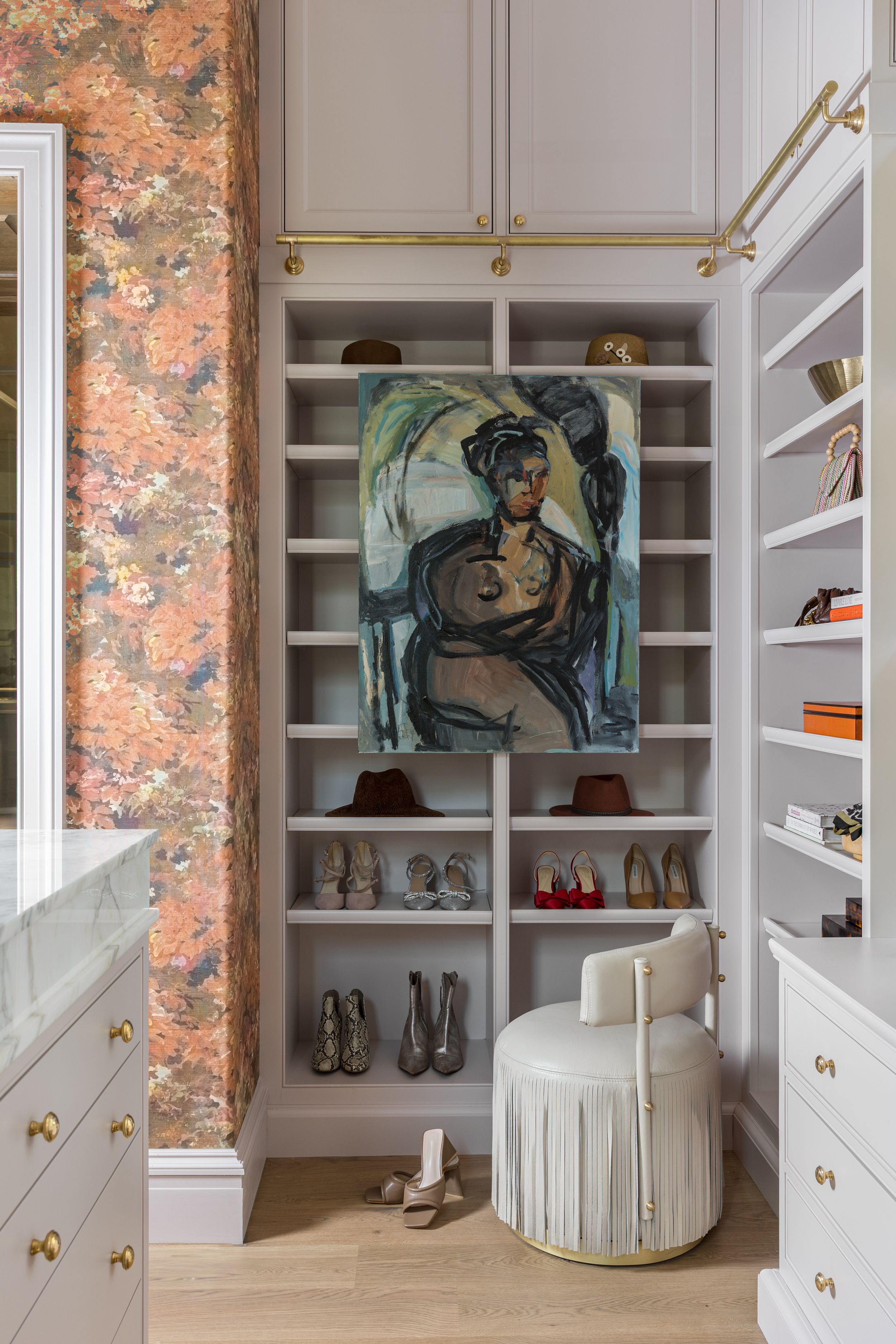A luxurious walk-in closet features built-in white shelving, floral wallpaper, brass hardware, and an artistic portrait display.